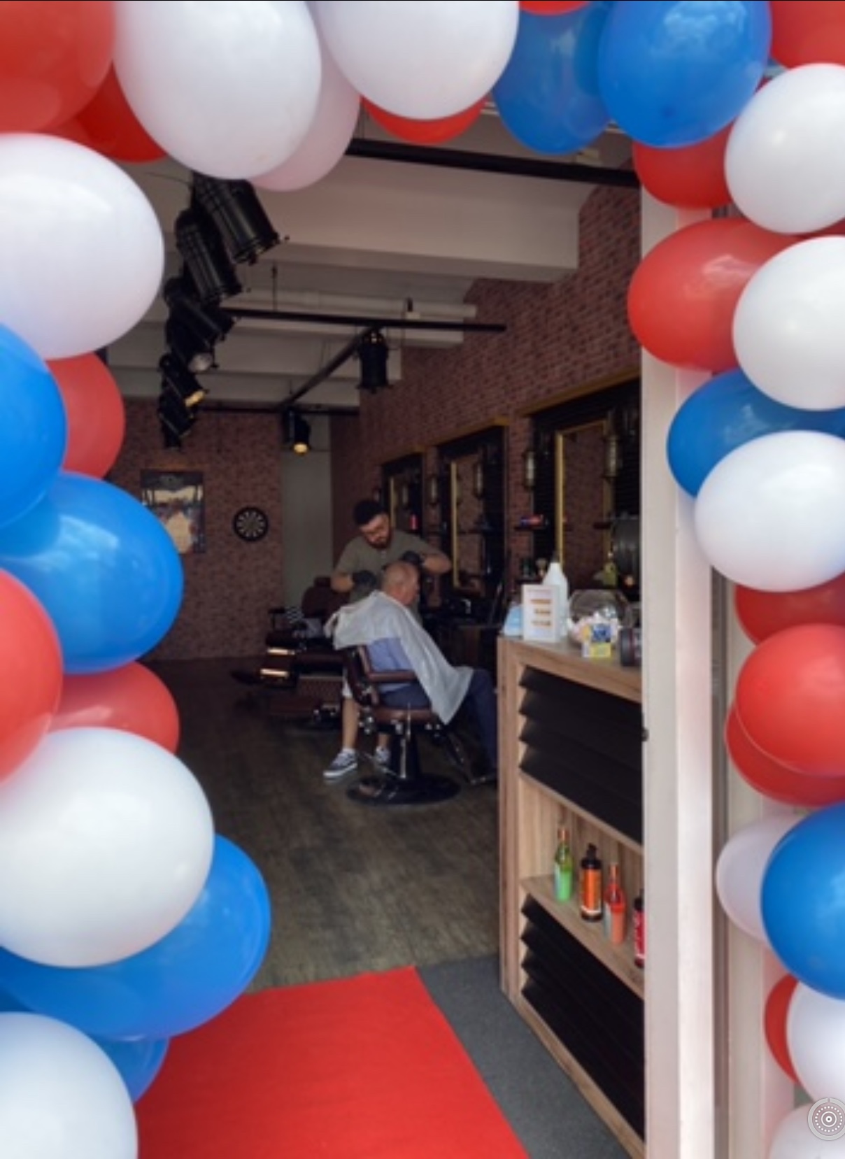 Doorkijk door een rood, wit en blauwe ballonnenboog de zaak in, met een klant in de stoel